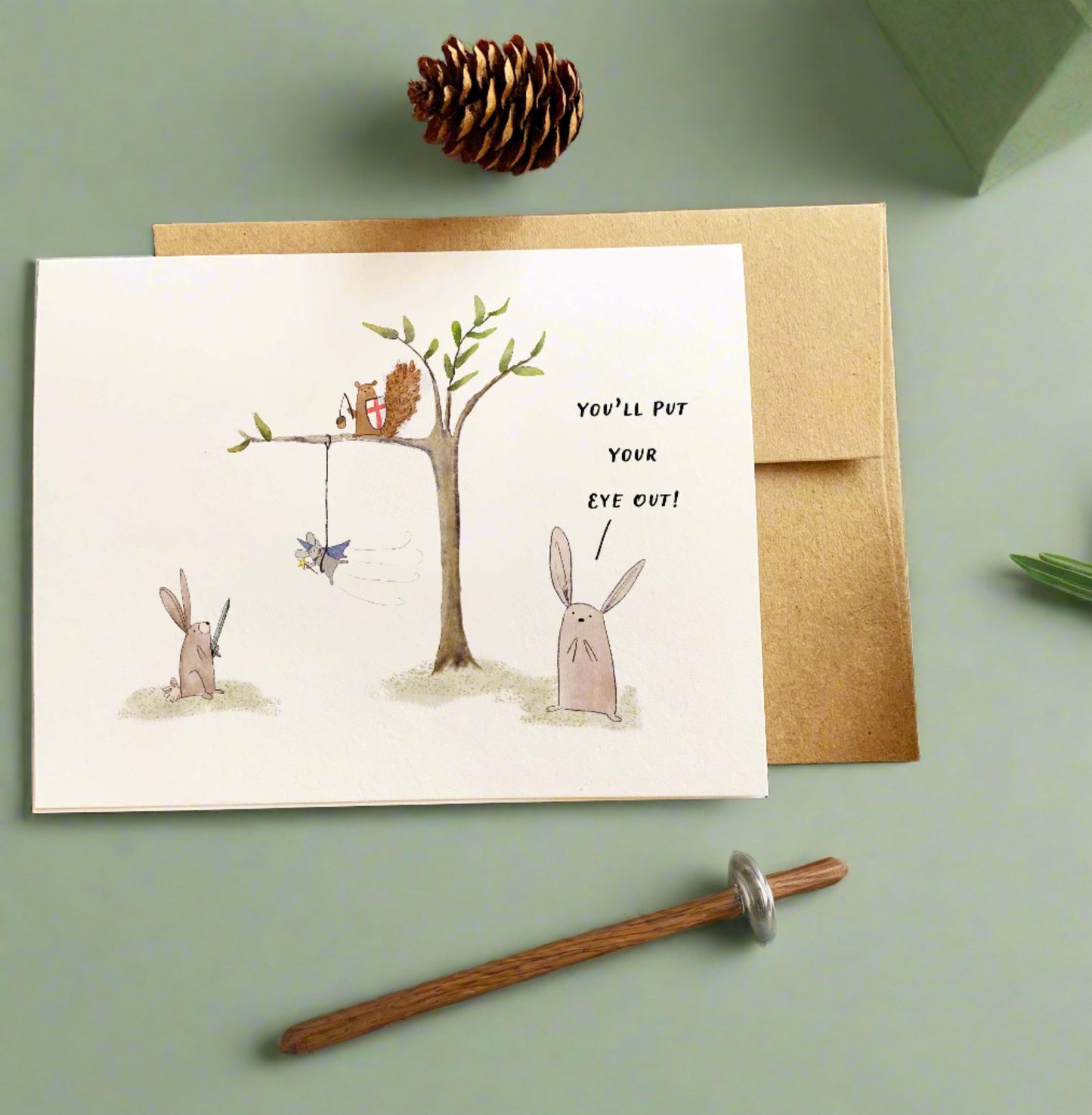 Some woodland friends are playing under a tree. A bunny holding a toy sword looks up into a tree where a mouse dressed as a wizard swings from a branch; just above is a squirrel holding a shield and acorn flail. A rabbit looks on, concerned, with the phrase "YOU'LL PUT YOUR EYE OUT!" written above.