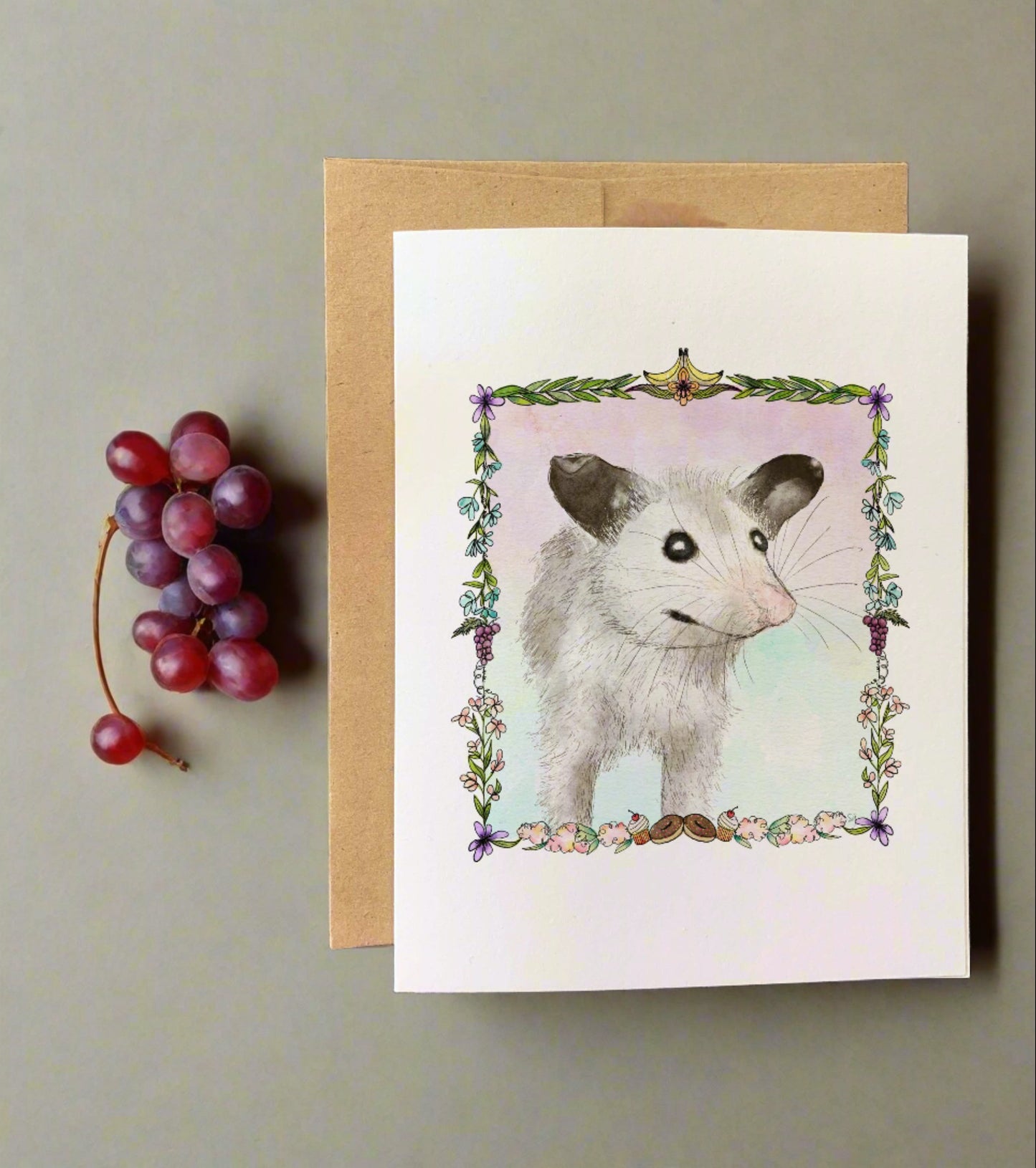 A greeting card with an opossum framed by its favortie snacks of fruit, donuts, bananas and cupcakes is bathed in soft light, a bunch of grapes sits next to it waiting to be eaten.